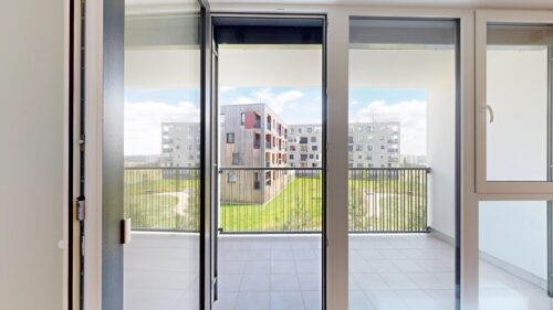 Sliding glass balcony doors open to a view of a modern apartment complex with a green lawn outside, under a blue sky.