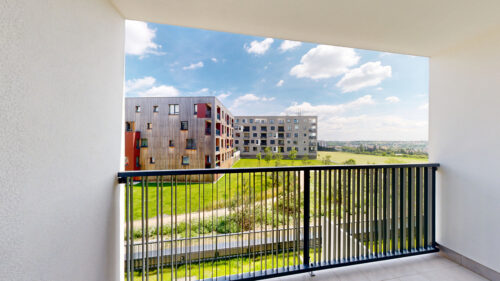 Balcony railing frames a view of modern apartment buildings with a green lawn and blue sky above.