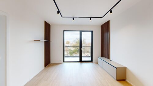 Bright empty living room with a large glass door to a balcony, white walls, light wood flooring, and vertical wooden slats on both sides outside the door frame and window view.