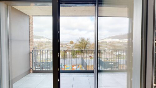 Interior view of a glass sliding door opening to a balcony with a metal railing and cityscape beyond.