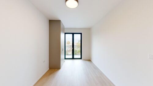 Bright empty hallway room with built-in brown wardrobe along left wall and a large glass door at the far end leading to a balcony, white walls, and light wood flooring.