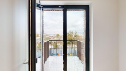 Open glass door onto a small balcony with a metal railing and city view beyond.”] ,