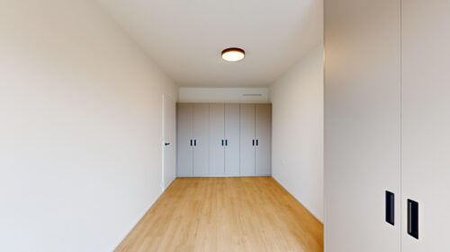 Narrow hallway with light wood floor and a row of gray cabinet doors along the back wall, white walls, and a circular ceiling light.