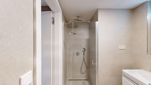 Modern bathroom with a glass shower enclosure, rainfall showerhead, handheld shower, beige tiled walls, and a white vanity.