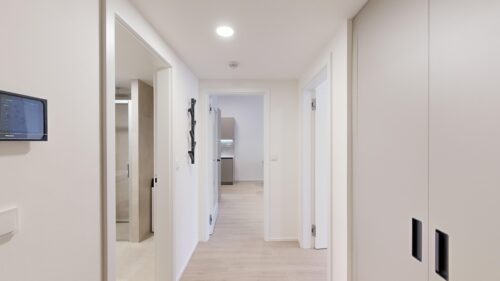 Bright, modern hallway with white walls, light wood floor, and doors along the corridor leading to a kitchen at the end.