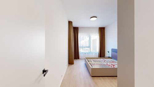 Minimal bedroom with a light wood floor, white walls, and a large window with sheer white curtains and brown drapes. A beige bed frame is visible near the window, with a small bedside lamp on a table to the right.