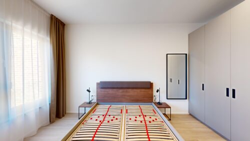 Modern bedroom with a wooden bed frame, two nightstands, and black desk lamps facing a large built-in wardrobe; sheer curtains by a window on the left.
