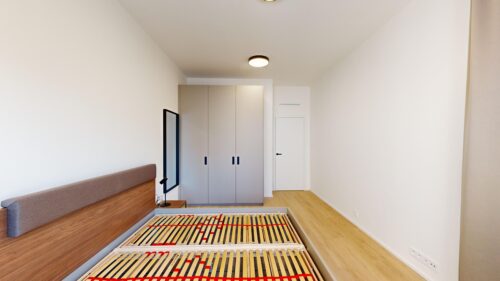 A modern bedroom with a wooden bed frame and upholstered headboard, a large gray wardrobe, and a ceiling light on white walls.