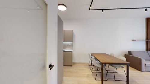Modern open-plan dining area with a wooden table and white chairs, a gray kitchen in the back, and a brown sofa to the right.
