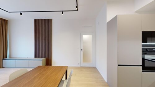 Open-plan room with a wooden dining table in the foreground, white walls, and built-in kitchen cabinets on the right, plus a door with frosted glass.