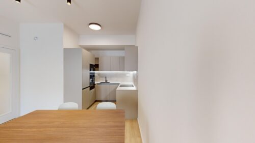 Modern kitchen seen through an opening from a dining area, with gray cabinets, built-in oven, and sink; wooden table in the foreground.