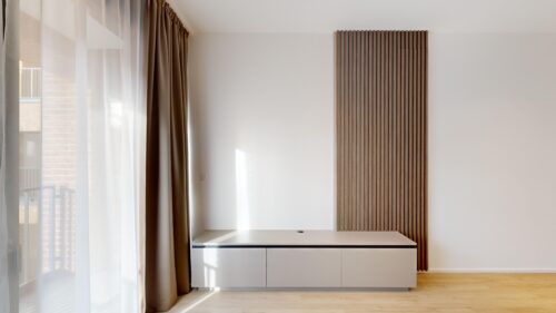 Minimalist living area with a white cabinet, vertical wood slat wall accent, and sheer curtains by a large window.