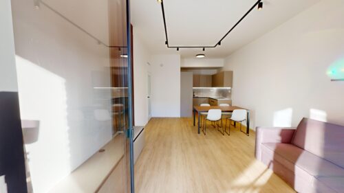 Bright open-plan kitchen and dining area: wooden dining table with white chairs, beige cabinetry, and track lighting along the ceiling. A pink sofa is visible in the foreground on the right.