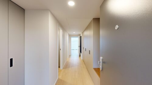 Bright, modern interior hallway with white walls, light wood flooring, and built-in storage along the right, illuminated by ceiling lights into the distance.