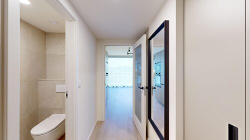 Modern hallway with white walls and open doors leading to a bright living area; bathroom visible on the left with a toilet and tiled wall.