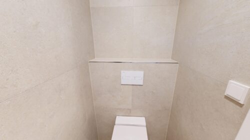 Narrow beige tiled bathroom with a wall-mounted toilet and a rectangular flush plate on the wall behind it.