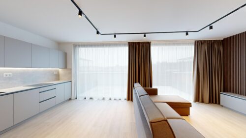Modern kitchen and living area with grey cabinets, light countertops, and track lighting in a bright space with sheer white curtains and brown drapes.