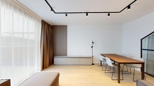 Bright minimalist living/dining room with a wooden table and white chairs by large windows with sheer white curtains and brown drapes.