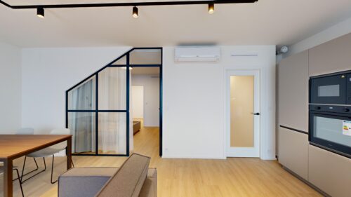 Modern open-plan living area with a glass-framed partition to a bedroom, white walls, and warm wood flooring.