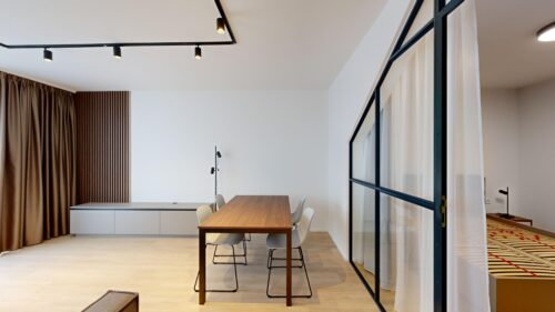 Modern conference room with a wooden table, white chairs, and a black-framed glass divider along the right; brown curtains on the left.
