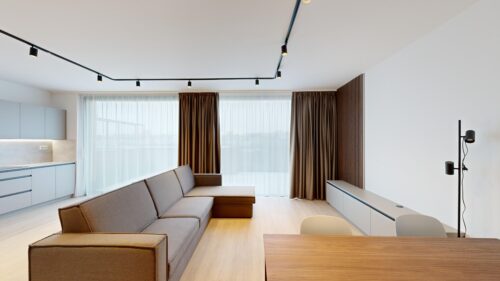 Bright modern living room with an L-shaped gray sofa, light wood floors, and white cabinets along the left wall. Large windows with sheer white curtains and brown drapes dominate the back wall, plus a black track lighting system overhead and a tall floor lamp on the right side.
