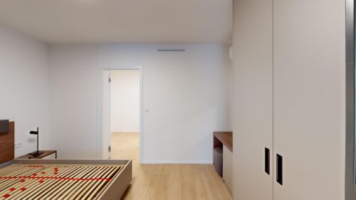 Bedroom with a partially assembled bed frame, a desk lamp on a side table, and light wood flooring; tall white wardrobes line the right wall, with an open doorway to another room in the back.