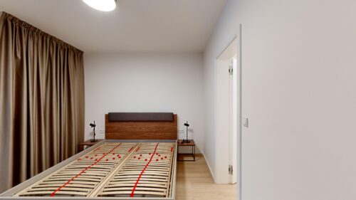 A minimalist bedroom with an open bed frame, two matching bedside tables, and a brown curtain along the left wall under a ceiling light.