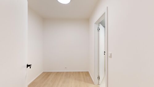 Bright hallway with light wood floor, white walls, a partially open white door on the right, and a round ceiling light.