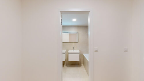 Through a doorway, a modern beige-tiled bathroom with a white sink, toilet, bathtub, and rectangular backlit mirror.