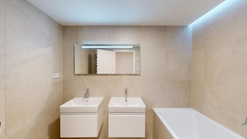 Modern bathroom with two white rectangular sinks and a wide mirror above, beige tiled walls, and a bathtub on the right edge.