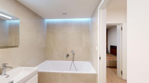 Modern bathroom with a white bathtub, beige tiled walls, and chrome fixtures; doorway to another room visible beyond the tub.