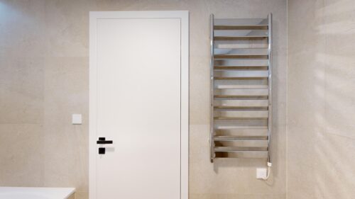 White interior door with black handle beside a chrome towel warmer mounted on a beige tiled wall in a bathroom layout.