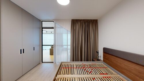 Modern bedroom with gray built-in wardrobes on the left, a wooden bed frame with slats in the foreground, and sheer white plus brown curtains over a glass door to a balcony.