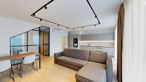 Open-concept living area with a brown sectional sofa, wooden dining table, and a modern kitchen along the back wall; glass partition with blue frames on the left.