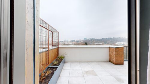 Rooftop balcony with gray tiled floor, metal grid railing, and planter boxes overlooking a cityscape under an overcast sky