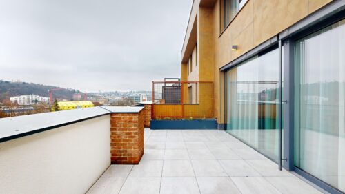 Rooftop terrace of a modern building with glass doors, brick planters, and a distant cityscape under a cloudy sky.