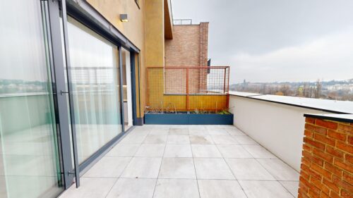 Balcony terrace with sliding glass doors, metal railing, and a long planter box overlooking a city under a cloudy sky.