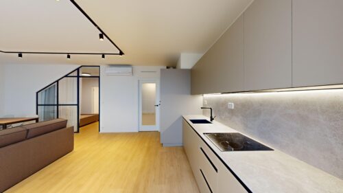 Open-plan kitchen and living area with grey upper cabinets, light countertops, and a black induction cooktop on the right.