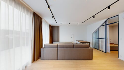 Open-plan living area with a beige sectional sofa, light wood floor, and floor-to-ceiling white and brown curtains along large windows.
