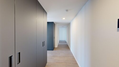 Long hallway with gray built-in wardrobes on the left, white walls, and a light wood floor, ending in a doorway.