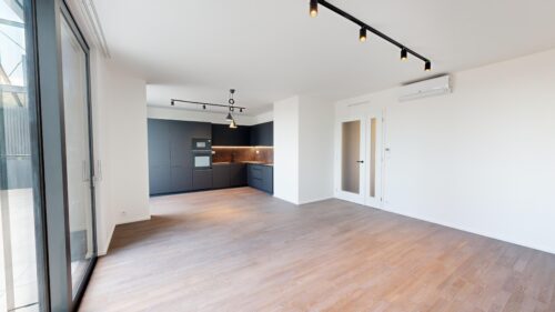 Open-plan kitchen area with dark gray cabinets, built-in oven, and a brown backsplash under black track lighting on white walls.