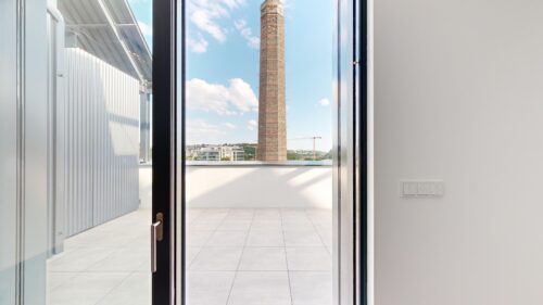 Interior doorway opens to a tiled balcony; a tall brick chimney rises outside with a construction crane in the distance under a blue sky.