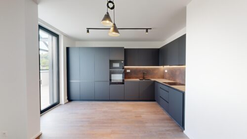Modern dark gray kitchen with tall matte cabinets, built-in oven and microwave, and warm under-cabinet lighting on a brown backsplashGenre.