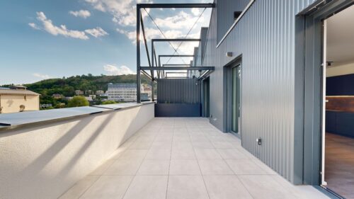 Rooftop terrace of a modern building with pale tiles, blue-gray corrugated walls, and exposed steel framing; hills and city in the distance.
