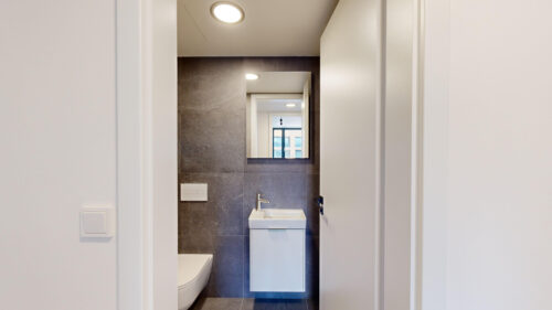 Narrow bathroom with dark gray tile wall, white sink and wall-mounted toilet, and a rectangular mirror reflecting a window.