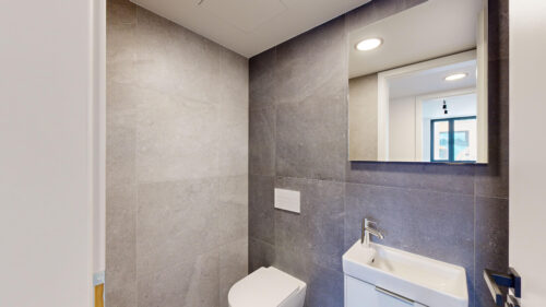 Modern bathroom with gray wall tiles, a white sink, wall-mounted toilet, and a rectangular mirror reflecting a window.