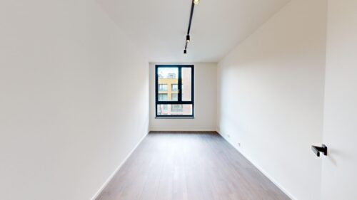 Empty narrow room with white walls, light wood floor, and a window at the far end showing a building exterior. A door handle is visible on the right side.