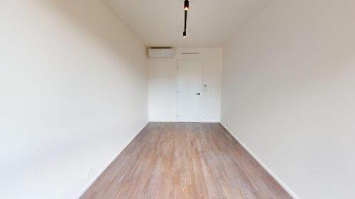 Narrow empty room with white walls, light wood flooring, a white door at the far end, and a ceiling track light above.
