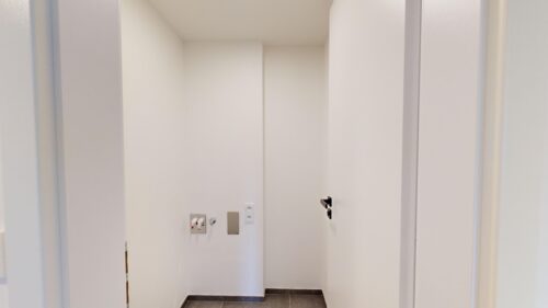 Narrow utility alcove with white walls, showing a small plumbing panel and electrical outlets near the back wall and a dark tile floor.