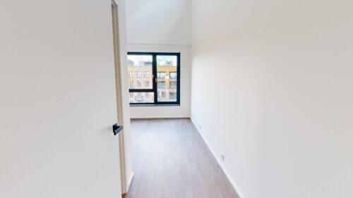 Narrow white-walled hallway with light wood floor leading to a large black-framed window showing brick buildings outside